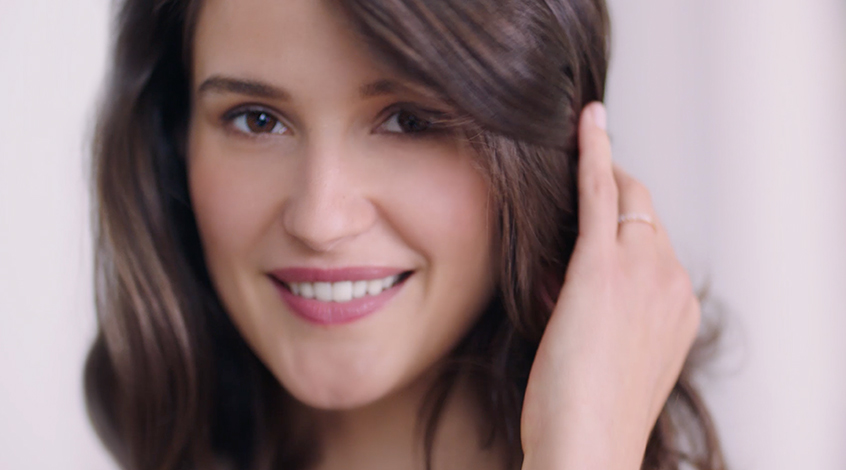 Woman with brown hair and brown eyes smiles into the camera while holding her hair back