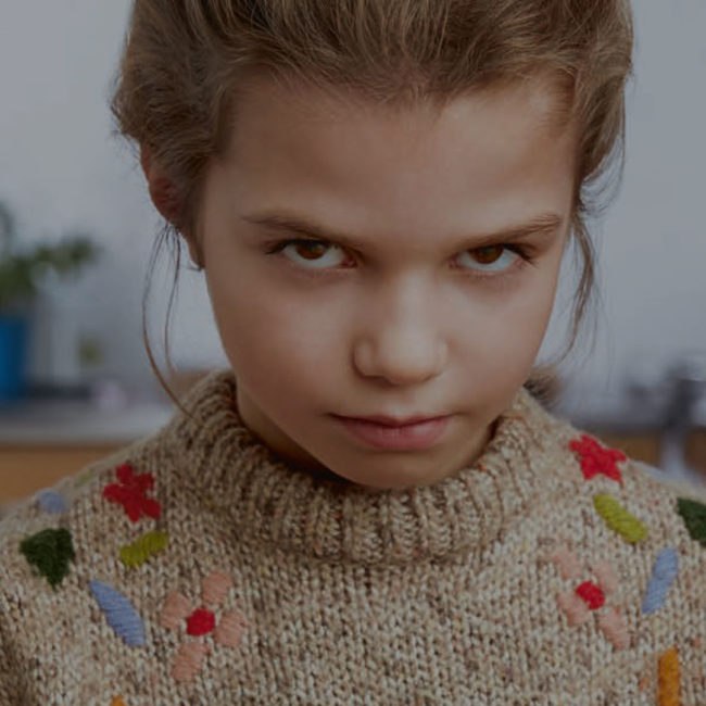 Mädchen mit braunen Augen und Strickpulli guckt schmollend in Kamera