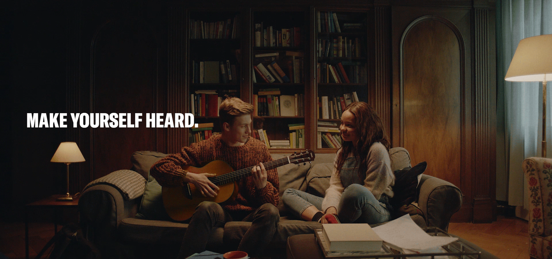 Boy plays guitar while sitting on the sofa with a girl, they both look at each other and smile, text "make yourself heared"